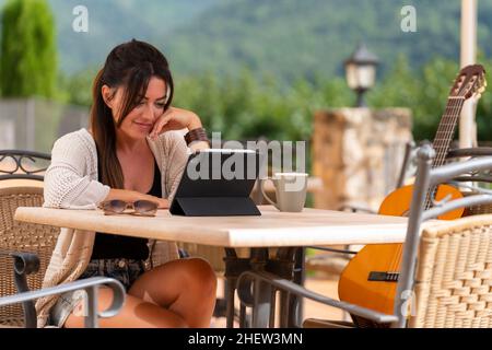 Frau, die auf einer Terrasse sitzt und mit ihrer spanischen Gitarre auf einem Stuhl auf das Tablet schaut Stockfoto