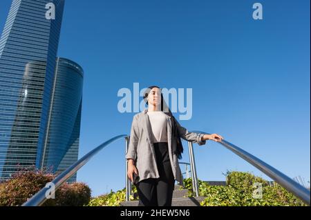 Aus dem Winkel betrachtet, wie eine Erwachsene Frau mit einem schwarzen Kopftuch auf einer Stufe einer Parktreppe steht und in einem sonnigen Bild die Kamera mit den Gebäuden im Hintergrund anschaut Stockfoto