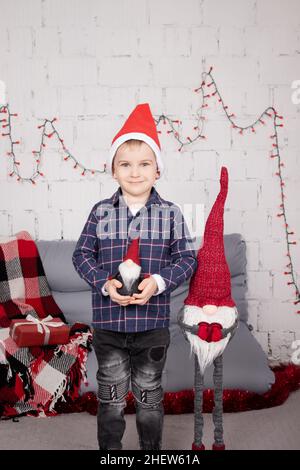 Kleiner Junge in santa hat in der Nähe weihnachten Gnom Spielzeug, halten Zwerg Spielzeug in der Hand in grauen Raum. weihnachtsdekor und Spielzeug. Kinder spielen. Stockfoto