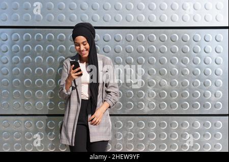 Vorderansicht einer fröhlichen muslimischen Frau, die mit einer Wand aus Eisenplatten mit Kreismustern hinter ihrem Telefon schaute und ihr Telefon benutzte. Stockfoto