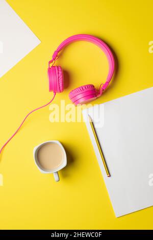 Rosafarbene kabelgebundene Kopfhörer mit mehreren leeren Papierblättern, einem Bleistift und einem Kaffee auf gelbem Hintergrund Stockfoto