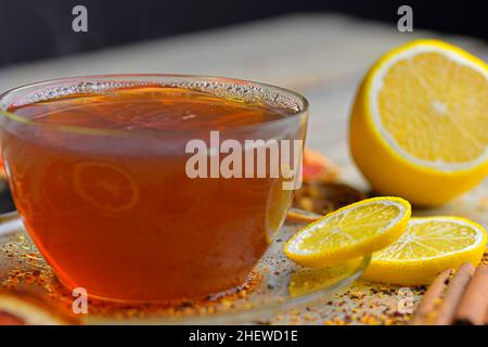 Glas Tasse Tee mit Zimtstangen auf Holztisch Hintergrund. Zimtschnecken, Sternanise, Zitrone und Trockenfrüchte. Schwarzer Hintergrund. Makrobild Stockfoto