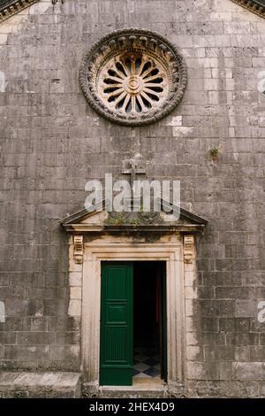 Rosenfenster mit Ornamenten an der dunklen Steinwand der Kirche über der grünen Holztür Stockfoto