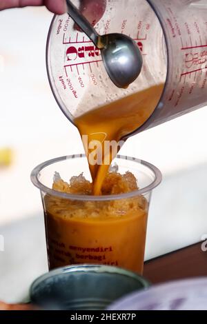 Gießen Sie einen frischen Thai-Tee aus brauner Milch in einer Messbecher in ein transparentes Kunststoffglas mit kaltrasiertem Eis im Inneren mit verschwommenem und weichem Fokus-Backgroun Stockfoto