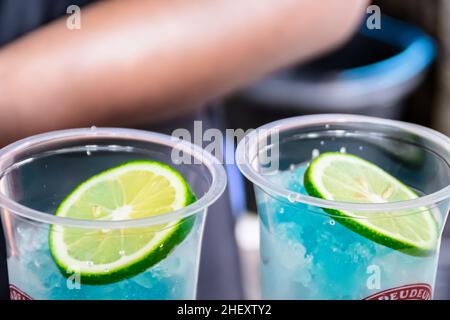 Kaltrasierte Eiswürfel mit gegossenem blauem Sirup und frischen sauren Limetten, die oben für einen Mojito-Drink in transparentem Kunststoffglas geschnitten wurden Stockfoto