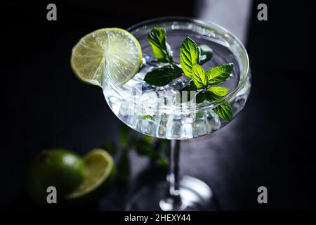 Gin Tonic im Cocktailglas mit Eis, Minzblättern und Limette auf einem dunklen rustikalen Brett Stockfoto