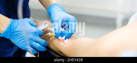 Die Tropfernadel in den Arm des Patienten einführen. Selektiver Fokus. Die Hände des Arztes in sterilen Handschuhen installieren einen Katheter zur intravenösen Verabreichung von Medikamenten an einen Patienten. Breitbild. Stockfoto