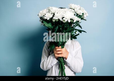 Ein nicht erkennbarer Junge in einem weißen Hemd hält einen Strauß weißer Blumen in seinen Händen und bedeckt sein Gesicht mit Blumen. Foto auf blauem Hintergrund. Motte Stockfoto