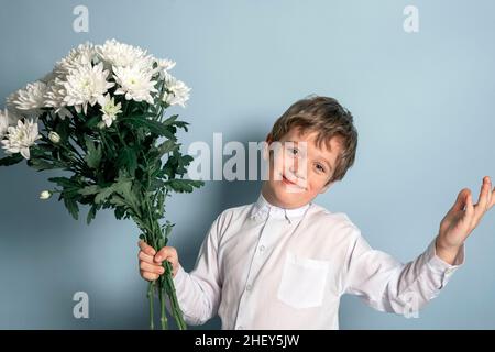 Ein süßer kaukasischer Junge in einem weißen Hemd hält einen Strauß weißer Blumen in der Hand und lächelt. Foto auf blauem Hintergrund. Muttertag, herzlichen Glückwunsch Stockfoto