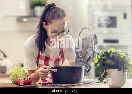 Hausfrau in Schürze öffnet Deckel eines Topfes, der in sie schaut, während sie in der Küche zu Hause kocht. Stockfoto