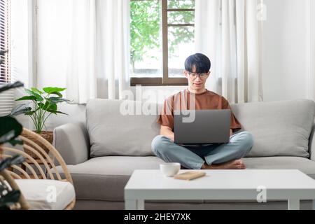 Technologiekonzept der Mann, der in legerer Kleidung auf dem Sofa sitzt, sein Notebook auf dem Schoß auslaht und seine Arbeit verrichten will. Stockfoto