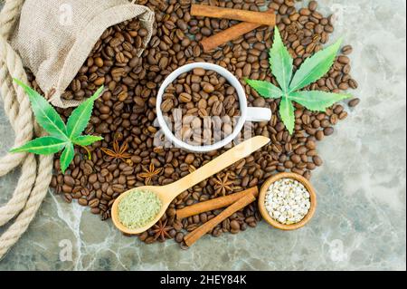 Eine Komposition aus Kaffeebohnen, einer Tasse gefüllt mit duftenden Kaffeebohnen, Cannabissamen und Morijuana-Blättern auf einem Marmorboden Stockfoto