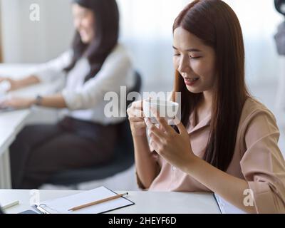 Asiatische junge weibliche Angestellte, die an ihrem Schreibtisch heißen Tee nippte. Geschäftsfrau entspannte sich vor der Arbeit mit einem heißen Getränk. Stockfoto