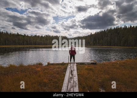 Der Entdecker und Reiseliebhaber in einem rot-schwarzen Hemd und einer grauen Mütze steht am Rande eines Holzwegs und genießt den Blick auf den Palolampi-See in HII Stockfoto