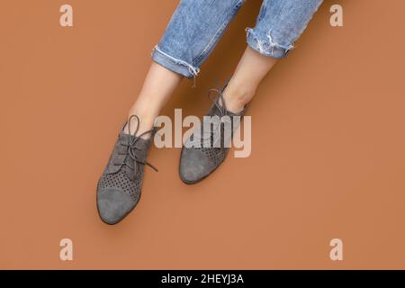 Die kaukasischen Beine der Frau in blau gedrehten Jeans und grauen Wildlederschuhen aus englischer Brogue Oxfords auf braunem beigen Hintergrund isoliert. Lässige Sneaker. Oberteil Stockfoto