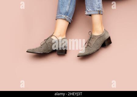 Die kaukasischen Beine der Frau sind in blau gedrehten Jeans und in grauem Wildleder gehaltene Schuhe aus englischer Brogue Oxfords auf hellrosa beigem Hintergrund isoliert. Lässige Sneaker. Fro Stockfoto