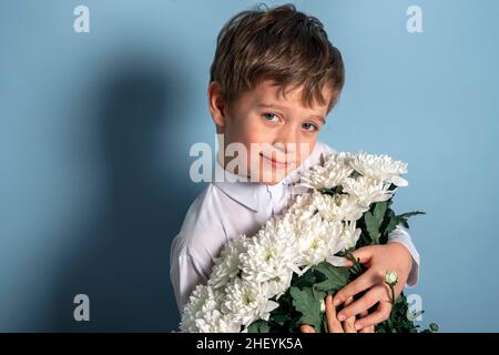 Ein süßer kaukasischer Junge in einem weißen Hemd hält einen Strauß weißer Blumen in seinen Händen und lächelt. Foto auf blauem Hintergrund. Muttertag, herzlichen Glückwunsch Stockfoto