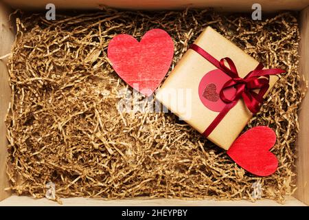 Holzkiste mit Geschenk in braunem Bastelpapier mit rotem Band eingewickelt, gefüllt mit Papierfüller, Draufsicht von oben Stockfoto