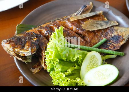 Gegrillter Karpfen in Sojasauce mit frischem grünem Gemüse und Gurke auf einem braunen Teller in einem Restaurant. Stockfoto