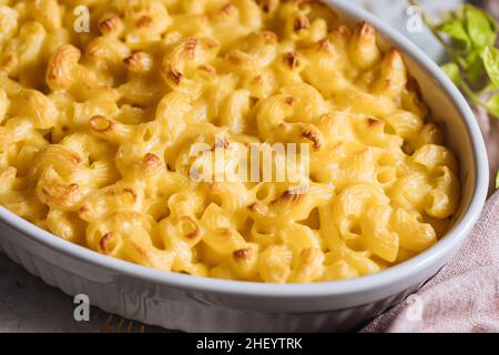 Klassischer mac und Käse mit Basilikum auf der Oberseite und mac und Käse im Ofen im Hintergrund mit Gabeln und rohen Makkaroni auf einem rustikalen Holzhintergrund gebacken Stockfoto