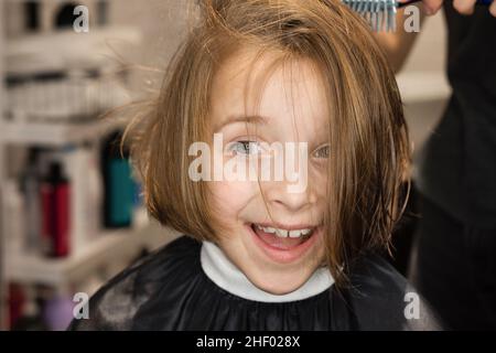 Angenehm überrascht, dass das Kind im Schönheitssalon einen Haarschnitt bekommen hat. Friseur mit Trockner nass Schuss Haar im Friseursalon. Kleines Mädchen mit dunkelblondem bob beim Stylist. Stil und Mode für Kinder. Stockfoto