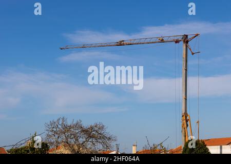 Ein Turmdrehkran, der auf einer Baustelle über den Dächern kleiner Wohnhäuser auf dem Land aufgestellt wurde, und ein blauer Himmel im Hintergrund Stockfoto