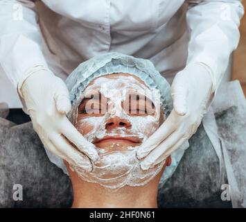 Kosmetikerin Anwendung Peeling-Maske auf das Gesicht zu jungen Mann, Spa Schönheitssalon. Das Konzept der Körper- und Gesichtspflege für Männer Stockfoto