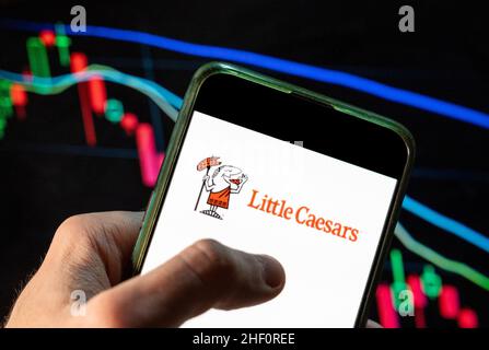 Auf dieser Fotolabbildung ist das Logo der amerikanischen drittgrößten Pizza-Restaurantkette Little Caesars zu sehen, das auf einem Smartphone mit einem Index-Diagramm der wirtschaftlichen Börse im Hintergrund zu sehen ist. Stockfoto