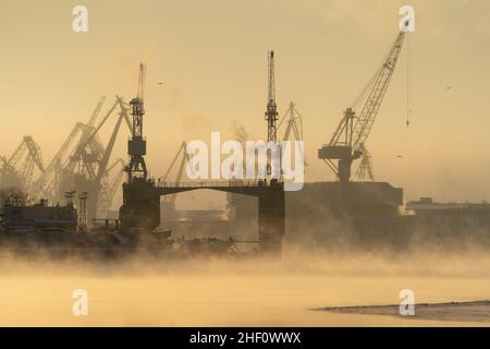 Kränen der Werft in frostigen Wintertag, Dampf über dem Fluss, glatte Oberfläche des Flusses bei Sonnenuntergang. Stockfoto