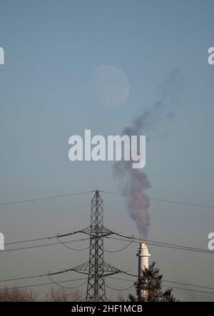 13. Januar 2022. Aufsteigender, tagsüber wachender, wuchtiger Mond nahe dem Horizont, im Vordergrund Power Pylon und Fabrikschornstein mit Rauch gegen den blauen Himmel. Merton, London, Großbritannien Stockfoto