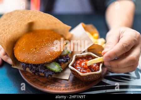Fastfood-Restaurant Hamburger und Pommes. Nahaufnahme eines Mannes, der Pommes frites in Tomatensauce eintaucht. Horizontale Aufnahme. Snack im Fast Food Restaurant Stockfoto