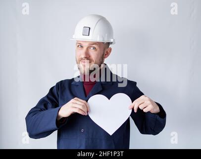Valentinstag: Männlicher Techniker, Auftragnehmer, Supervisor oder Ingenieur in Hardhat und blauer Uniform mit weißem Papier in Herzform. Romantik oder Beziehungskonzept. Hochwertige Bilder Stockfoto