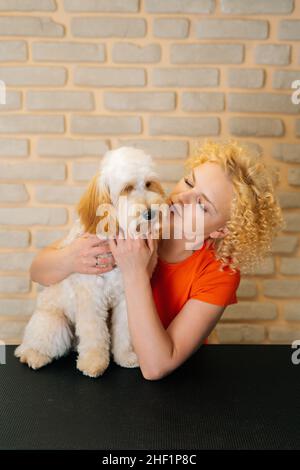Vertikales Porträt einer glücklichen weiblichen Groomerin, die einen lockigen Labradoodle-Hund nach dem Haarschnitt im Friseursalon hält. Stockfoto