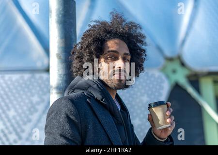 Schöner moderner Afro-Mann mit lächelndem Bart, der auf der Straße steht und eine Tasse Kaffee zum Mitnehmen trinkt Stockfoto