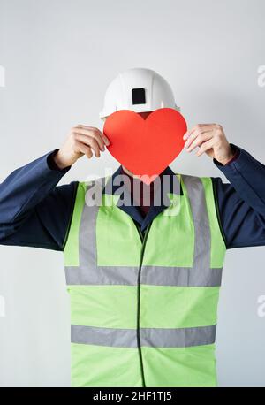 Porträt eines bärtigen Bauunternehmers, Vorgesetzten oder Ingenieurs in Helm, der an der Wand steht und vor dem Gesicht eine rote Herzform hält. Valentinstag-Konzept. Hochwertiges vertikales Bild Stockfoto