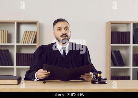 Junger Richter in einem Kleid sitzt an seinem Tisch im Gerichtssaal und liest den Satz Stockfoto