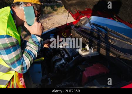 Kaukasischer Mann, der unter der Haube eines kaputten Autos mit einem Mechaniker telefoniert. Stockfoto