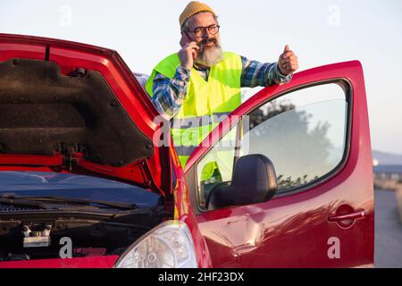 Erwachsener Mann, der ruhig aussaund am Telefon am Straßenrand telefoniert. Latino-Mann mit geöffneter Haube aufgrund eines mechanischen Problems Stockfoto