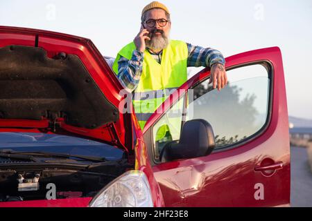 Erwachsener Mann, der ruhig aussaund am Telefon am Straßenrand telefoniert. Latino-Mann mit geöffneter Haube aufgrund eines mechanischen Problems Stockfoto