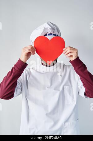 Kaukasischer bärtiger Koch oder Bäcker in weißer Uniform und rotem Papierherz vor dem Gesicht als St. Valentinstag-Konzept. Ein rotes Herz vor die Arbeitskleidung halten. Hochwertige Fotos Stockfoto
