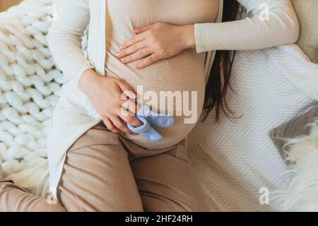 Mutterschafts- und Schwangerschaftskonzept. Beschnittenes Foto einer Schwangeren, die ein Paar niedliche blaue Babysocken für ein ungeborenes Kind auf dem Bauch hält, während sie sich zu Hause entspannt Stockfoto