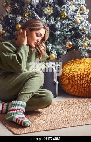 Winterurlaub. Gemütliches Hausfoto von jungen entspannten Frau in der Nähe geschmückten Weihnachtsbaum zu Hause, weiblich in bequemen Homewear und gestrickte Socken re Stockfoto