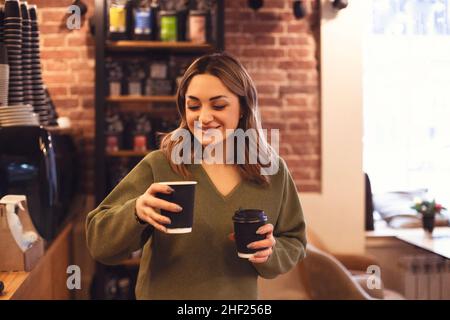 Innenfoto einer jungen lächelnden Frau, die zwei Kaffeetassen zum Mitnehmen hält, während sie im Coffeeshop oder Café steht, selektiver Fokus der Frau, die heiße drin bringt Stockfoto