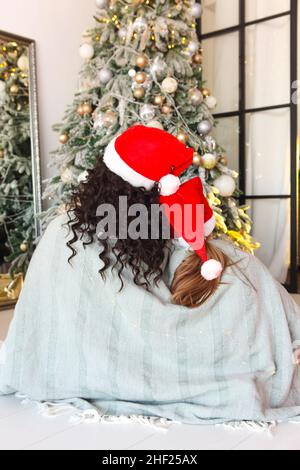 Weihnachten Für Die Ganze Familie. Rückansicht von Mutter und Kind mit weihnachtsmützen, die unter einer gestrickten Decke auf dem Boden sitzen und sich umarmen und auf den geschmückten Weihnachtsbaum blicken. Stockfoto