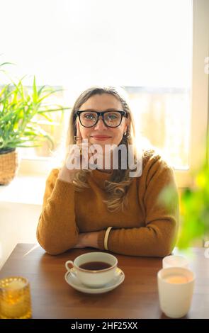 Porträt einer Frau mittleren Alters mit einer Tasse Tee, die mit brennenden Kerzen am Tisch sitzt, einer Frau mit einem heißen Morgengetränk, während sie am Holzschreibtisch sitzt Stockfoto