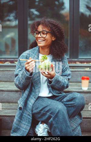 Glückliche afroamerikanische Büroangestellte, die Salat isst und die Kamera lächelt, während sie im Park auf der Bank sitzt, selektiver Fokus. Positiv schwarz Stockfoto