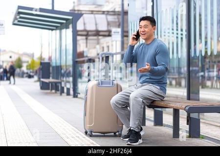 Tourist an der Bushaltestelle, Spaß am Telefon zu haben, auf einer Bank mit einem großen Koffer auf ein Taxi warten, Asian Mann auf einer Geschäftsreise Stockfoto