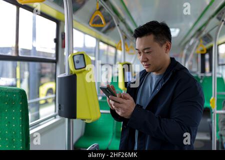 Glücklicher und erfolgreicher asiatischer männlicher Fahrgast im öffentlichen Verkehr kaufte ein Ticket mit einem lächelnden Handy und schaute auf die Kamera Stockfoto