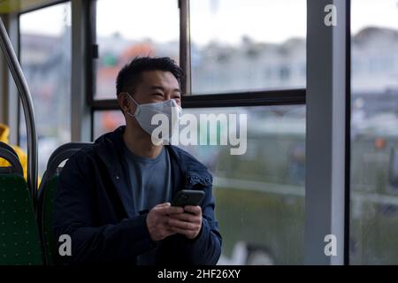 Ein asiatischer Passagier in einer schützenden medizinischen Maske auf seinem Gesicht schreibt und liest besorgt Nachrichten von einem Mobiltelefon, ein Mann reist durch die Stadt mit dem Publ Stockfoto