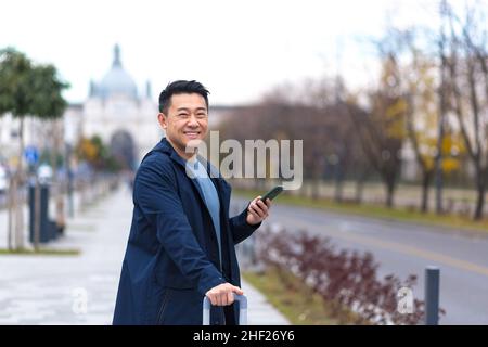 Fröhlicher asiatischer Tourist, warm angezogen, in einer europäischen Stadt, schaut auf die Kamera und lächelt, nutzt die Anwendung auf dem Telefon, um durch die Stadt zu navigieren Stockfoto